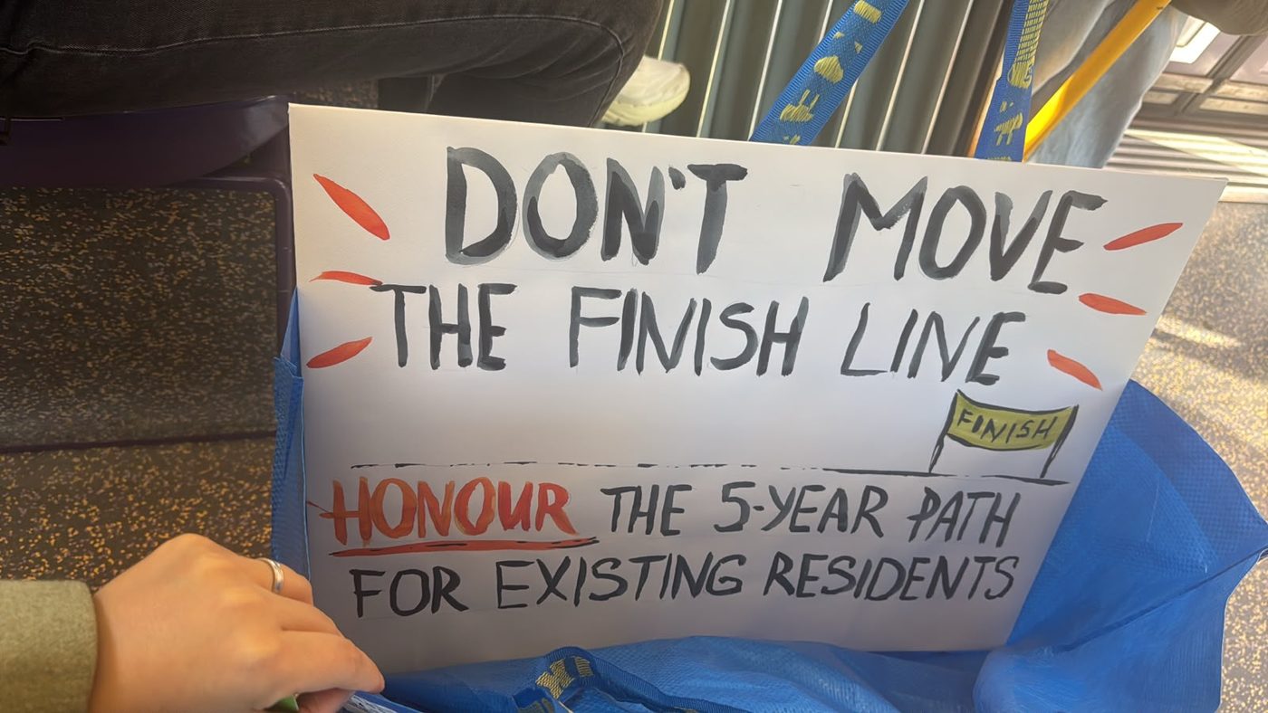 A handmade sign reading ‘Don’t move the finish line — honour the 5-year path for existing residents’
